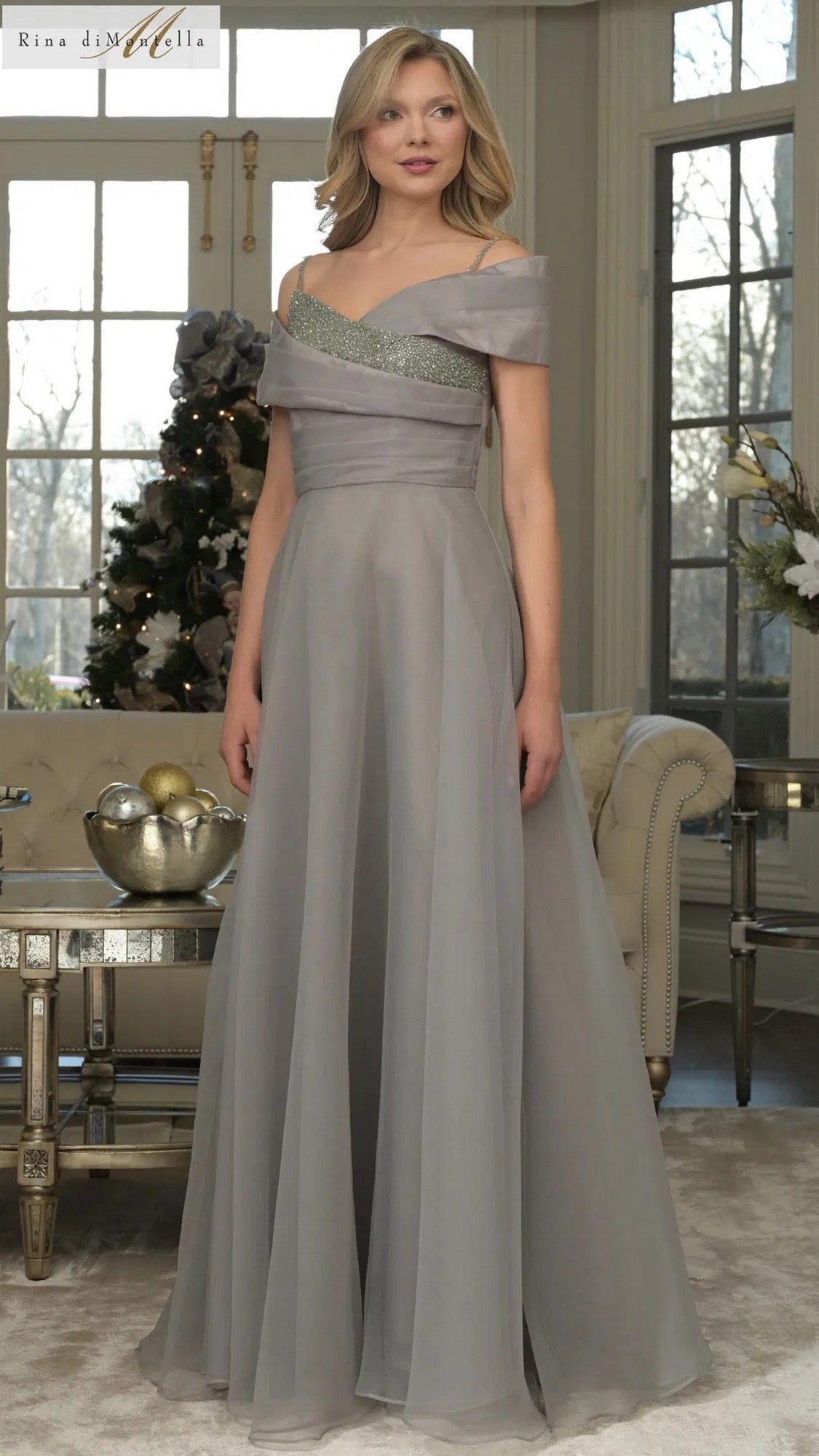 A woman stands indoors wearing the Rina Di Montella RD3303 Long Formal Dress in thyme, featuring a floor-length, off-shoulder neckline with a fitted bodice and flowing A-line skirt.